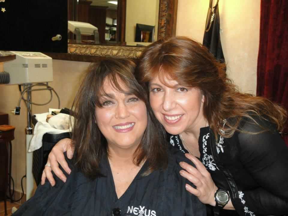 Two women smiling in a hair salon, one sitting and one standing behind her with a hand on her shoulder.